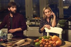 a man and woman sitting at a kitchen table talking on their cell phones, with oranges in the foreground