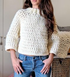 a woman standing in front of a bed wearing a white crochet sweater and jeans