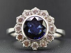 An incredibly glamorous flower adorns your finger in this impressive Ceylon Sapphire and sparkling Diamond Daisy cluster ring.  This daring yet feminine riff on the halo ring is one sparkling blossom that will never wilt or fade. Serving as the center of this bold flower ring is a striking two and a half carat Ceylon Sapphire, measuring 8mm in diameter.  This lively rich royal blue round shape Sapphire instantly grabs the eye with its saturated sparkle, communicating undeniable sophistication. T Engagement Dress, Ceylon Sapphire, Jewelry Workshop, Engagement Dresses, Two And A Half, Sparkling Diamond, Dress Rings, Platinum Ring, Halo Ring