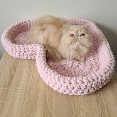 a cat sitting in a pink crocheted bed on top of a wooden table