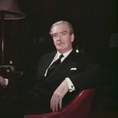 a man in a suit and tie sitting in a red chair next to a lamp