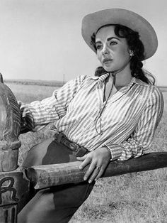 a woman wearing a cowboy hat leaning on a fence with a horse in the background