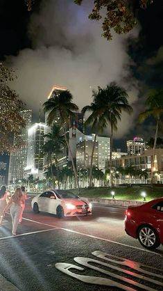 cars driving down the street at night with palm trees and buildings in the back ground