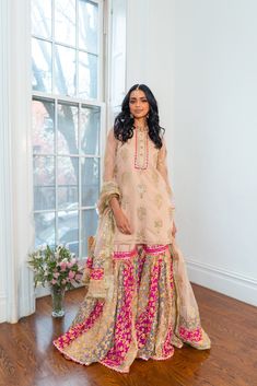 a woman standing in front of a window wearing a pink and gold outfit with matching dupatta