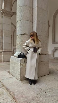 a woman leaning against a pillar wearing a long coat