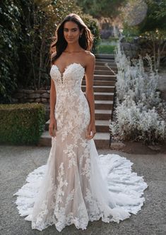 a woman in a white wedding dress standing on the ground next to some stairs and bushes