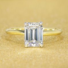 an emerald - cut diamond ring sits on a gold surface, with sparkle around it