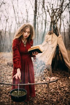 a woman in a red dress reading a book near a teepee and some trees