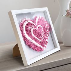 a pink heart in a white frame on a table next to a vase with flowers