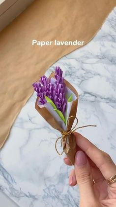 a hand holding a paper lavender flower in a brown box on a marble countertop