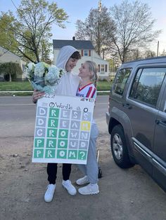 two people standing next to each other in front of a car holding flowers and a sign