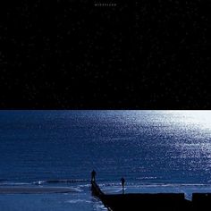 two people standing on the edge of a pier in the ocean at night with stars above