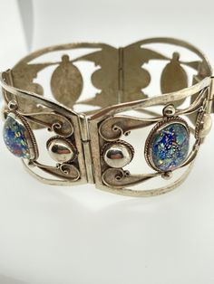 "Vintage Taxco Confetti Opal Glass & Sterling Silver Hinged Cutout Bracelet -Very unique intricate silver work with 4 hinged panels each holding one oval confetti glass piece -Bracelet is hallmarked \"Taxco Sterling 925\" -Push insert locking closure -Bracelet width measures 1.4\" -Bracelet interior diameter measures about 2.3\", circumference just over 7\" -Oval glass pieces each bezel set measure about 16mm x 12mm -Total weight 37.3g -In very good vintage condition with light patina to the sil Elegant Silver Bracelet With Glass, Elegant Silver Glass Bracelets, Party Jewelry In Silver With Glass Material, Silver Fusion Bracelets For Party, Silver Glass Jewelry For Party, Silver Glass Bracelets For Party, Silver Cabochon Jewelry For Party, Silver Oval Bracelets For Collectors, Silver Oval Bracelets With Intricate Design