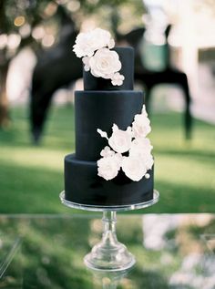 a three tiered cake with white flowers on top sitting on a glass table in front of a horse