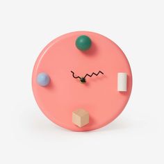 a pink clock with different colored balls and shapes on it's face, sitting against a white background