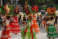 three women in colorful dresses and hats dance with their hands up while others watch from the sidelines