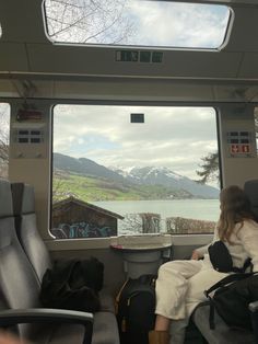 a woman sitting on a train looking out the window at mountains and water in the distance