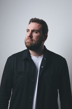 a man with a beard standing in front of a white wall and looking off to the side