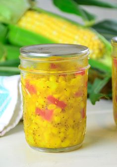 two jars filled with food sitting on top of a table next to some corn cobs