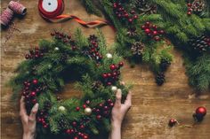 two hands are decorating a christmas wreath on a wooden table with pine cones, red berries and twine of twine