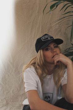 a woman sitting on the ground with her hand under her chin wearing a black hat