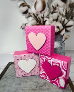 three pink boxes with hearts on them sitting next to a vase filled with cotton flowers