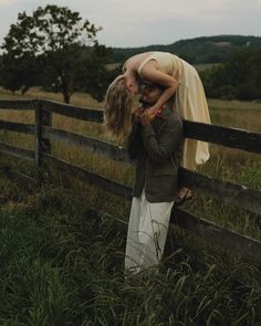 a man and woman kissing in front of a wooden fence on a grassy field with mountains in the background