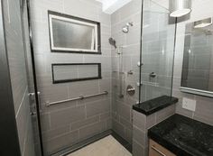 a walk in shower sitting next to a black counter top under a bathroom mirror and light