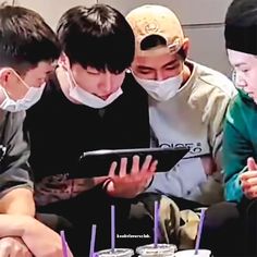 three boys wearing face masks are looking at an electronic device while another boy looks on