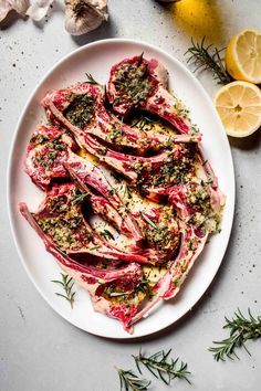 a white plate topped with meat covered in herbs and lemon wedges next to garlic