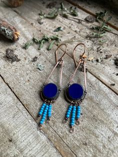 two blue beads are hanging from earrings on a wooden surface with leaves and twigs in the background