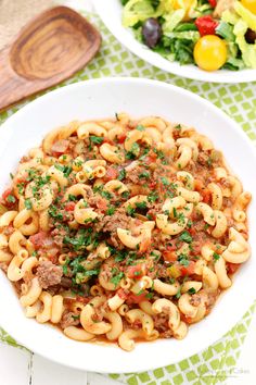 pasta with meat and vegetables in a white bowl on a green tablecloth next to a salad