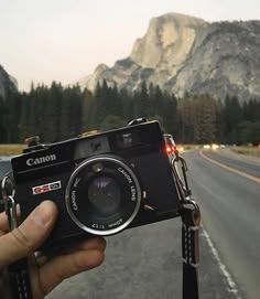 a person holding up a camera in front of a road with mountains and trees behind them