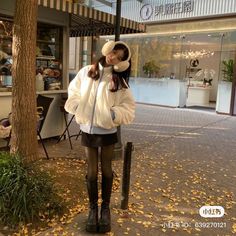 a woman standing on the sidewalk in front of a tree wearing a white jacket and black boots