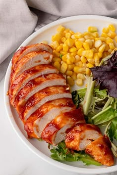 a white plate topped with meat, corn and lettuce next to a salad