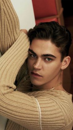 a young man in a tan sweater leaning against a wall with his hands on his head