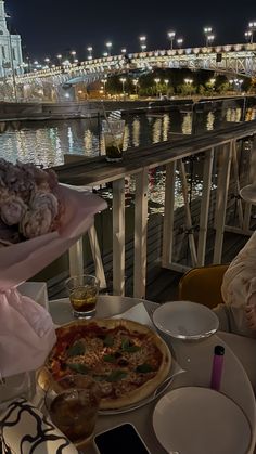 two people sitting at a table with pizza and drinks in front of them, overlooking the water