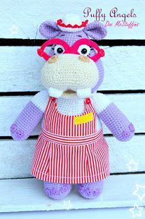 a crocheted stuffed animal wearing an apron and eyeglasses is posed on a white wooden surface