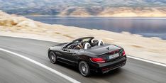 a black convertible car driving down a road next to the ocean and mountains in the background