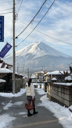 a woman walking down a snow covered street with a mountain in the background and power lines above her