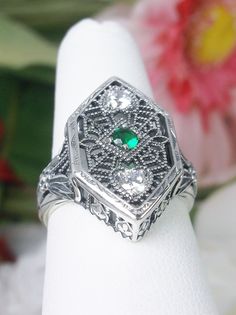 Simulated Green Emerald & White Cubic Zirconia (CZ) Sterling Silver RingCharlotte Design#D231 Resurrected from the Art Deco movement, this gorgeous Art Deco reproduction filigree ring is crafted in sterling silver. This lovely filigree ring is set with a stunning quality 2mm center round-cut simulated green emerald gemstone. Two small white cubic zirconia gemstones also grace the face of the ring, one near each point, each set in the center of a tiny heart. There are 2 filigree accent flowers th Heirloom Sterling Silver Diamond Ring With Filigree, Silver Art Deco Emerald Wedding Ring, Art Deco Silver Emerald Wedding Ring, Silver Emerald Ring With Intricate Design For Anniversary, Anniversary Silver Emerald Ring With Intricate Design, Vintage Silver Filigree Ring With Diamond Accents, Heirloom Emerald Ring With Intricate Design For Anniversary, Heirloom Sterling Silver Filigree Ring With Diamond Accents, Vintage Silver Emerald Ring With Diamond Accents