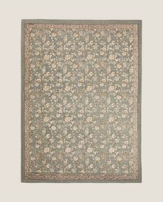 a blue rug with white flowers on the bottom and green border, in front of a gray wall