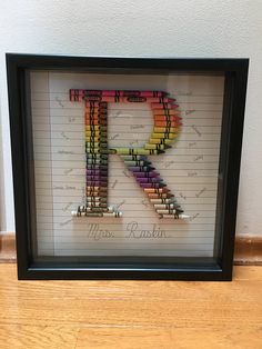 the letter k made out of crayons is displayed in a black frame on a wooden floor