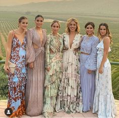 four women in dresses posing for a photo at an outdoor event with vineyards in the background