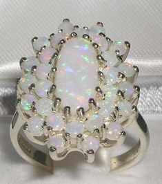 This large floral ring is made from 9K White Gold with Natural Semi-precious Opals.*Total gem weight : 7.26ctA extraordinary large 9ct Gold Fiery Opal Cluster ring set with a center large 12x6mm (0.48"x0.24" inches) complimented by fourteen 2.75mm (0.11" inches) and fourteen 2.25mm (0.09" inches) fiery Australian Opals. This large cluster ring is a real beauty and guaranteed to impress. TotalHeight:     8.15 millimetersWidth:  17 milliimetersLength:  18 milliimetersA rainbow of colors sparkle wi Unique White Ring With Accent Stones, Unique White Rings With Accent Stones, Dazzling White Cluster Ring Gift, White Gemstone Flower Ring, White Gemstone Cluster Ring As A Gift, Fine Jewelry White Flower Ring With Gemstone, Unique White Opal Gemstone Ring, White Cluster Jewelry With Halo Setting, Elegant White Flower Ring With Gemstone