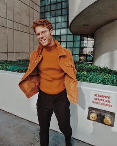 a man in an orange sweater and black pants standing next to a planter with plants