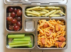 a lunch box filled with pasta, grapes and sausages next to celery