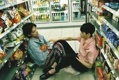 two people sitting on the floor in front of a store filled with food and drinks