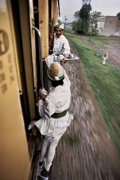 two men are riding on a train and one is holding a tray with food in it