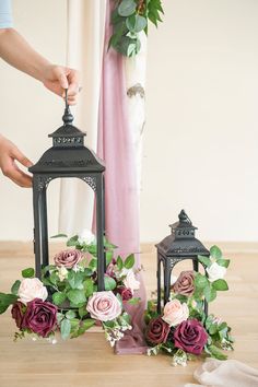 two black lanterns with pink and red flowers in them on a wooden table next to a curtain